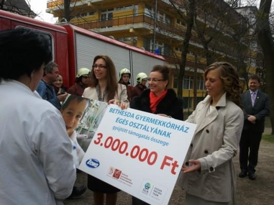 Az adományt jelképező csekket adja át a Kórház dolgozóinak (balról jobbra) Sándor Judit, kováts Adél és Jaksa Petra. Háttérben Rozgonyi Zoltán alpolgármester és a Zuglói Önkéntes Tűzoltók.